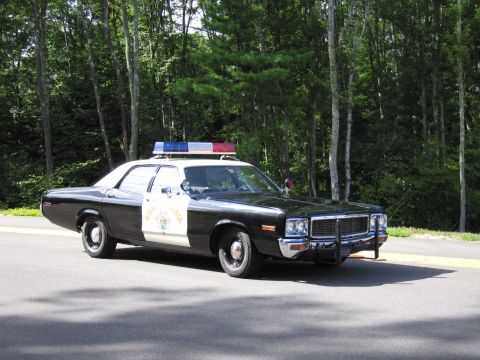 1991 Ford LTD Crown Victoria - black-and-white 1970s full-size police sedan marked Highway Patrol with a roof lightbar sits on a paved road in front of dense green trees.