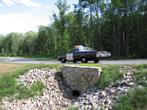 1991 Ford LTD Crown Victoria - black-and-white Highway Patrol police sedan with a roof lightbar parked along a road, seen behind a stone-faced drainage culvert and rocks.