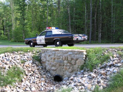1991 Ford LTD Crown Victoria - black-and-white Highway Patrol police sedan with star door emblem and roof lightbar parked by woods, above a stone culvert with rocks.