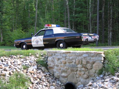 1991 Ford LTD Crown Victoria - black-and-white police sedan marked Highway Patrol with roof lights parked along a wooded road behind a fieldstone retaining wall.