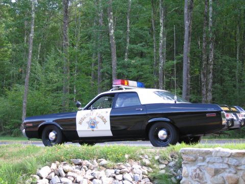 1991 Ford LTD Crown Victoria - Driver-side view of a black-and-white Highway Patrol police sedan with roof lightbar and tall antenna, parked beside a stone wall and fores…