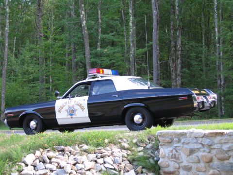 1991 Ford LTD Crown Victoria - black-and-white Highway Patrol police sedan with star door emblem and roof lightbar parked on a road by woods and a low stone wall.