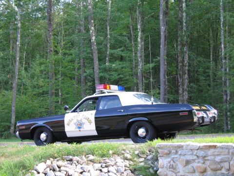1991 Ford LTD Crown Victoria - Driver-side profile of a black-and-white Highway Patrol police sedan with roof lightbar parked beside a stone wall and dense forest.