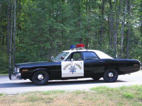 1991 Ford LTD Crown Victoria - Driver-side profile of a black-and-white Highway Patrol police sedan with roof lightbar and grille guard parked on a road beside green tree…