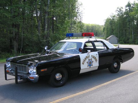 1991 Ford LTD Crown Victoria - Front three-quarter view of a black-and-white Highway Patrol police sedan with grille guard and roof lightbar parked on a road by woods.