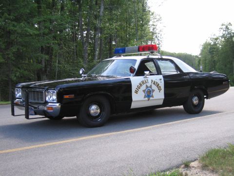 1991 Ford LTD Crown Victoria - Front three-quarter view of a black-and-white Highway Patrol police sedan with grille guard and roof lightbar on a wooded paved road.