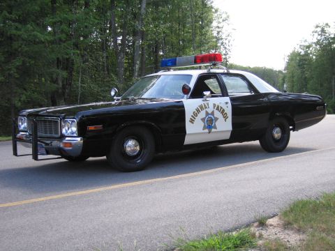 1991 Ford LTD Crown Victoria - black-and-white Highway Patrol police sedan with grille guard and roof lightbar parked at the edge of a wooded road.