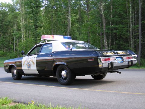 1991 Ford LTD Crown Victoria - Rear three-quarter view of a black-and-white Highway Patrol police sedan with chrome bumpers and roof lightbar parked on a wooded road.