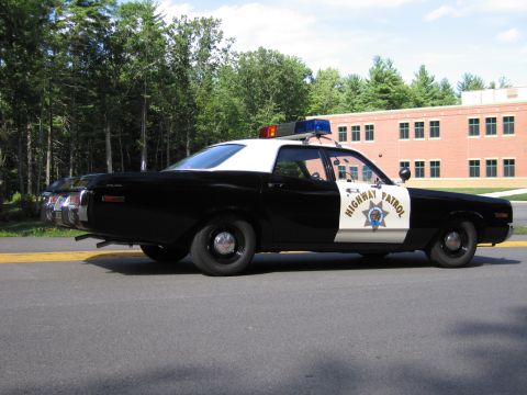 1991 Ford LTD Crown Victoria - Driver-side rear view of a black-and-white Highway Patrol police sedan with roof lightbar parked on a road by trees and a brick building.