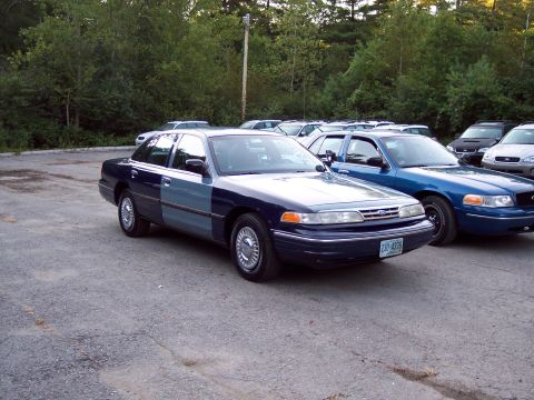1991 Ford LTD Crown Victoria - two-tone blue and silver Ford sedan parked at an angle in a gravel lot alongside other cars, with trees in the background.