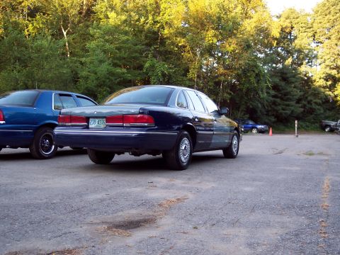 1991 Ford LTD Crown Victoria - Rear three-quarter view of a dark-blue Ford sedan parked beside a blue coupe in a gravel lot, with woods and a traffic cone behind.
