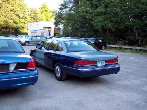 1991 Ford LTD Crown Victoria - Rear three-quarter view of a two-tone blue and gray Ford sedan parked next to a brighter blue Ford in a lot, with a building and trees behi…