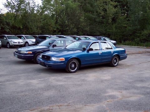 1991 Ford LTD Crown Victoria - bright blue Ford sedan and a darker teal Ford sedan parked side by side in a gravel lot with rows of vehicles and woods behind.