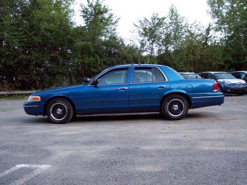 1991 Ford LTD Crown Victoria - teal-blue four-door sedan sits in profile on a gravel parking lot backed by green trees, with other cars in the distance.