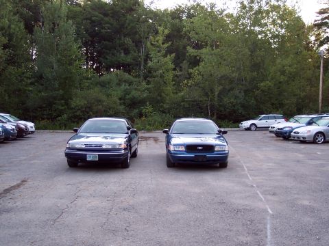 1991 Ford LTD Crown Victoria - dark blue Ford sedan and a brighter blue Ford sedan are parked nose-out side by side on a paved lot lined with trees.