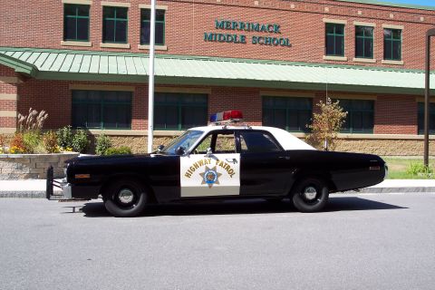 1991 Ford LTD Crown Victoria - black-and-white Highway Patrol sedan with a roof light bar is parked in profile outside a brick building lettered 'Merrimack Middle School.'