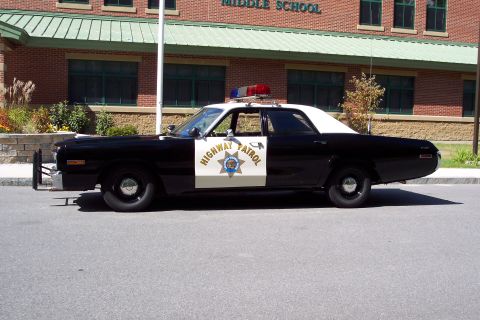 1991 Ford LTD Crown Victoria - Profile view of a black-and-white Highway Patrol sedan showing the star door decal, roof light bar, and a brick school building behind.