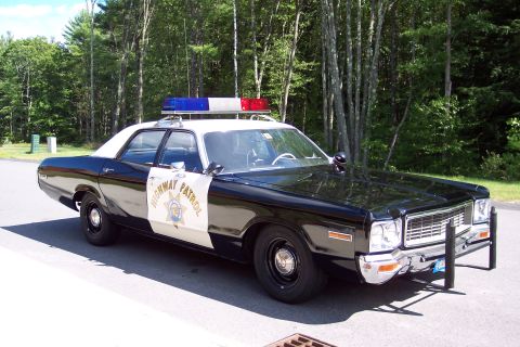 1991 Ford LTD Crown Victoria - Front three-quarter view of a black-and-white Highway Patrol sedan with chrome push bar and roof light bar on a road beside green woods.