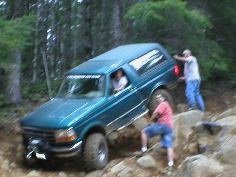 1978 Ford Bronco - A teal Ford Bronco navigates a challenging rocky trail with assistance from bystanders.