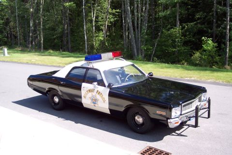 1991 Ford LTD Crown Victoria - black-and-white Highway Patrol sedan with push bar and roof light bar sits on shaded pavement with grass and trees behind.