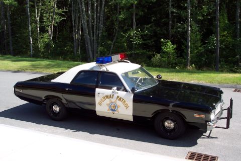 1991 Ford LTD Crown Victoria - Profile view of a black-and-white Highway Patrol sedan with roof light bar and star door decal parked on a road backed by green trees.