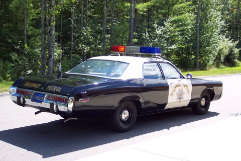 1991 Ford LTD Crown Victoria - Rear three-quarter view of a black-and-white Highway Patrol sedan with trunk and door 'HIGHWAY PATROL' lettering parked by green trees.