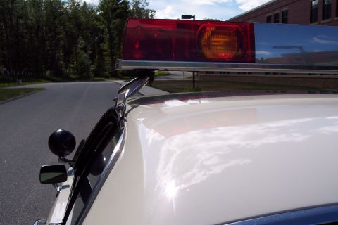 1991 Ford LTD Crown Victoria - Close-up of a red and amber roof light bar mounted above a white car's hood and chrome spotlight, with a road and trees behind.