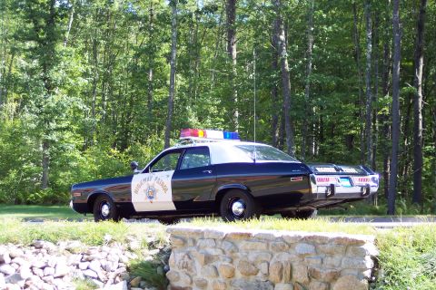 1991 Ford LTD Crown Victoria - Rear three-quarter view of a black-and-white Highway Patrol sedan with roof light bar and door star decal, parked beside a stone wall and t…
