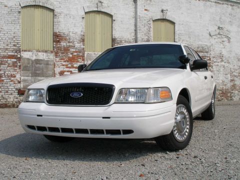 2001 Ford Crown Victoria - A white Ford Crown Victoria is parked on a gravel lot in front of a weathered brick building.