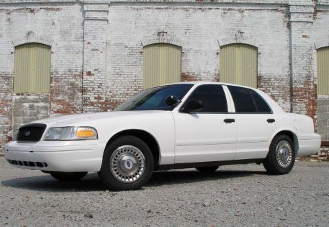 2001 Ford Crown Victoria - A white 2001 Ford Crown Victoria sedan is viewed from the front three-quarter angle, parked in front of a weathered brick building.