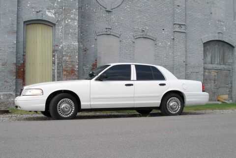 2001 Ford Crown Victoria - A white 2001 Ford Crown Victoria sedan is parked on a street next to a weathered brick building.