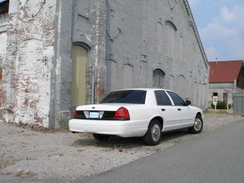 2001 Ford Crown Victoria - This white 2001 Ford Crown Victoria is parked on a gravel lot next to a large brick building.