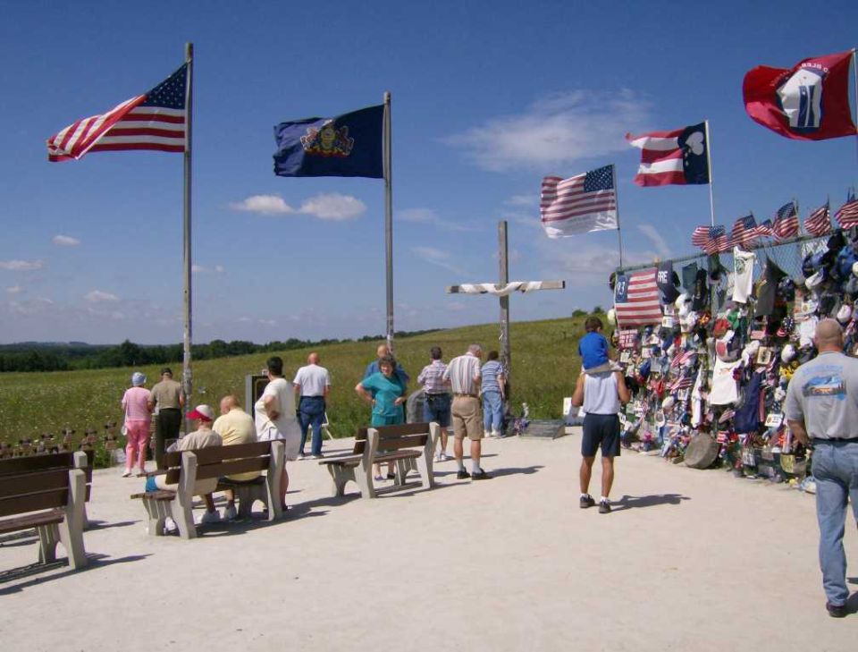 Memorial site with American and state flags, a cross, and a wall covered in photos and mementos