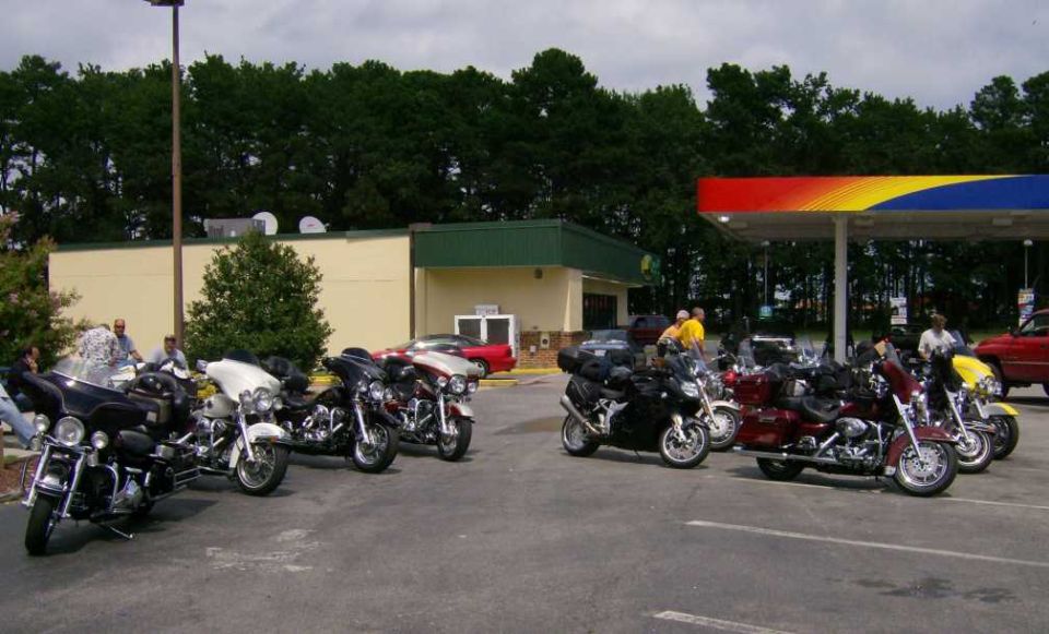 Several motorcycles parked in a lot near a gas station canopy. A red car is visible in the background