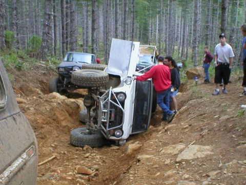 1978 Ford Bronco - A white off-road vehicle has flipped onto its side while navigating a rough trail, with a blue SUV visible behind it.