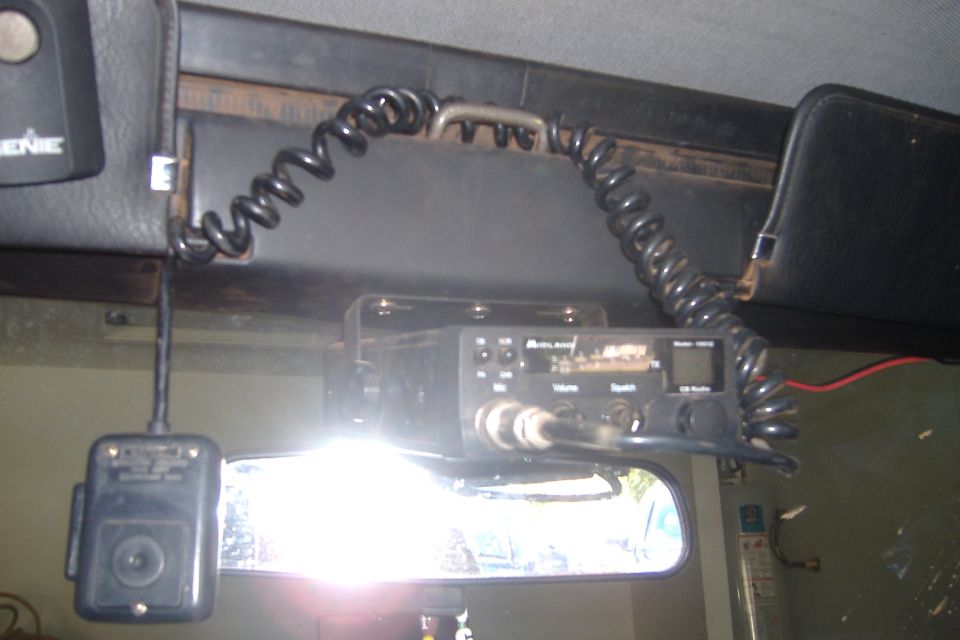 Midland CB radio mounted to a vehicle headliner with coiled mic cord and mirror dash cam