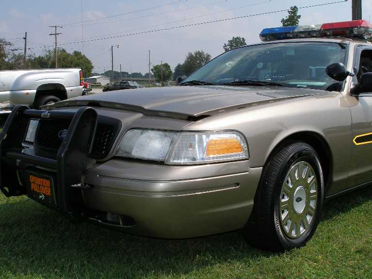 Tan Ford Crown Victoria police car with push bumper and roof light bar on grass