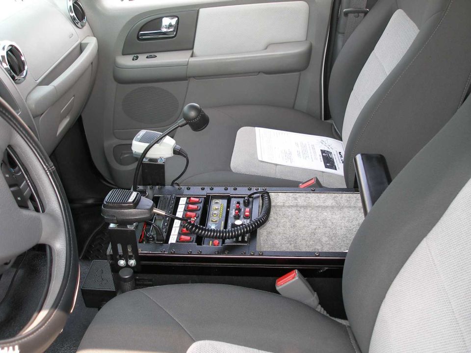 Vehicle interior with center-mounted police radio console, switches, mic, and gray cloth seats.