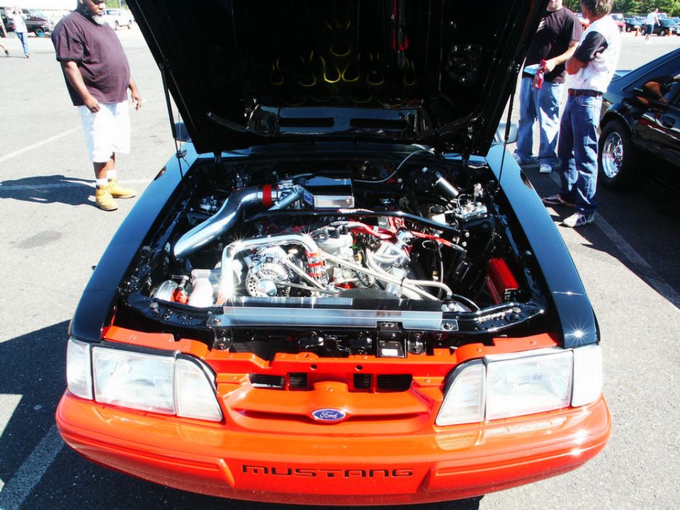 Front view of a modified Ford Mustang engine bay with the hood open. Orange front bumper with 'MUSTANG' text