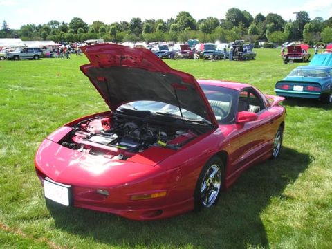 1984 Pontiac Fiero - A red 1994 Pontiac Firebird Trans Am is displayed with its hood open at a car show, with other vehicles and tents in the background.