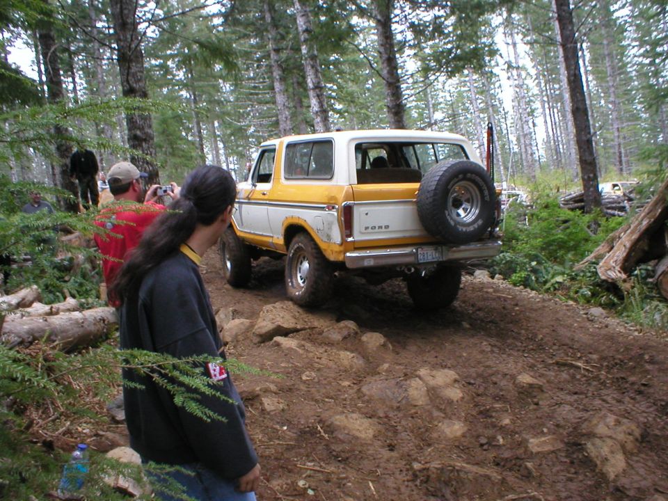 Rear of a yellow and white Ford Bronco with swing-out spare tire carrier on a rocky forest trail