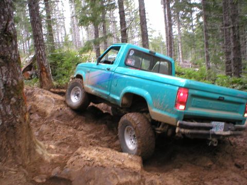 1978 Ford Bronco - A teal pickup truck navigates a steep, muddy trail surrounded by trees.