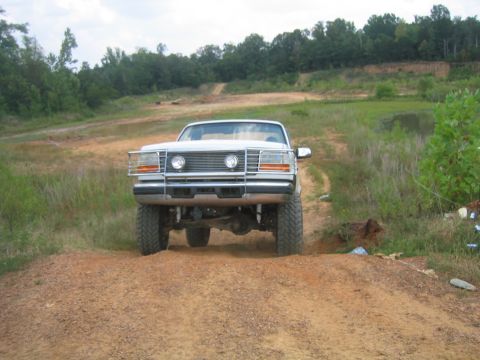 1996 Ford Bronco - A white Ford F-Series truck is shown driving on a dirt path with trees and a pond in the background.