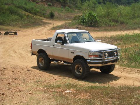 1996 Ford Bronco - This lifted white Ford Bronco is parked on a dirt road with a wooded hill in the background.