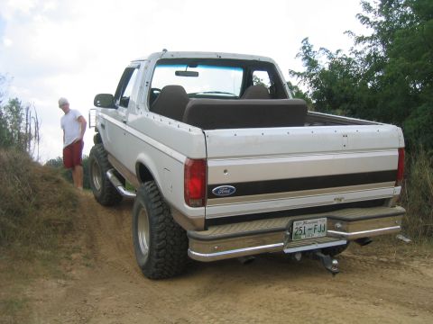 1996 Ford Bronco - A white 1996 Ford Bronco with lifted suspension and off-road tires is seen from the rear on a dirt path.