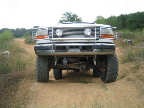 1996 Ford Bronco - A lifted white Ford Bronco is shown from the front on a dirt path, with trees in the background.