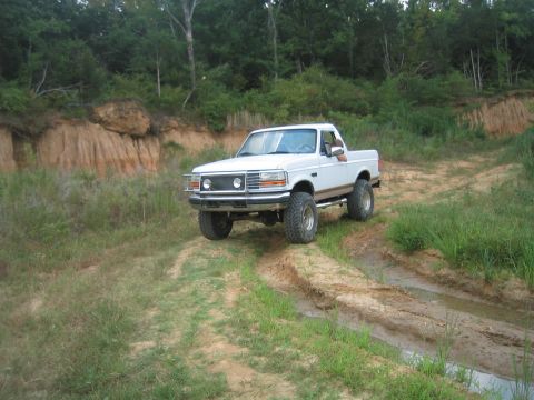 1996 Ford Bronco - White pickup truck navigates an off-road trail with muddy patches and grassy banks.