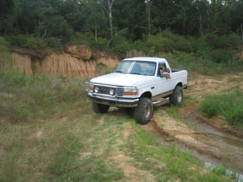 1996 Ford Bronco - This white pickup truck navigates a muddy dirt path surrounded by greenery.