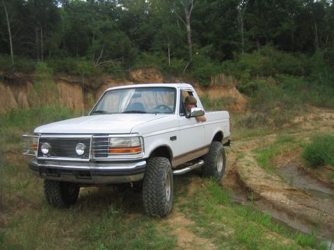 1996 Ford Bronco - White pickup truck with off-road tires driving on a dirt path through grassy terrain.
