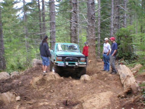 1978 Ford Bronco - A teal Ford Bronco navigates a muddy trail through a dense forest with several people observing.